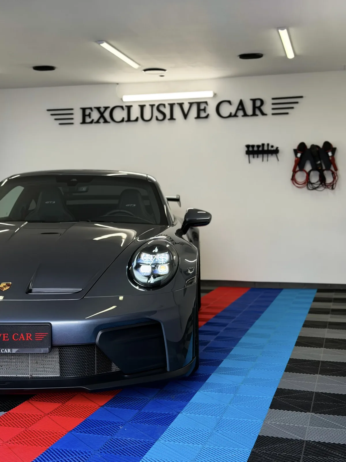 Šedé Porsche 911 GT3 v dílně Exclusive Car s logem na stěně a nářadím pro detailing
